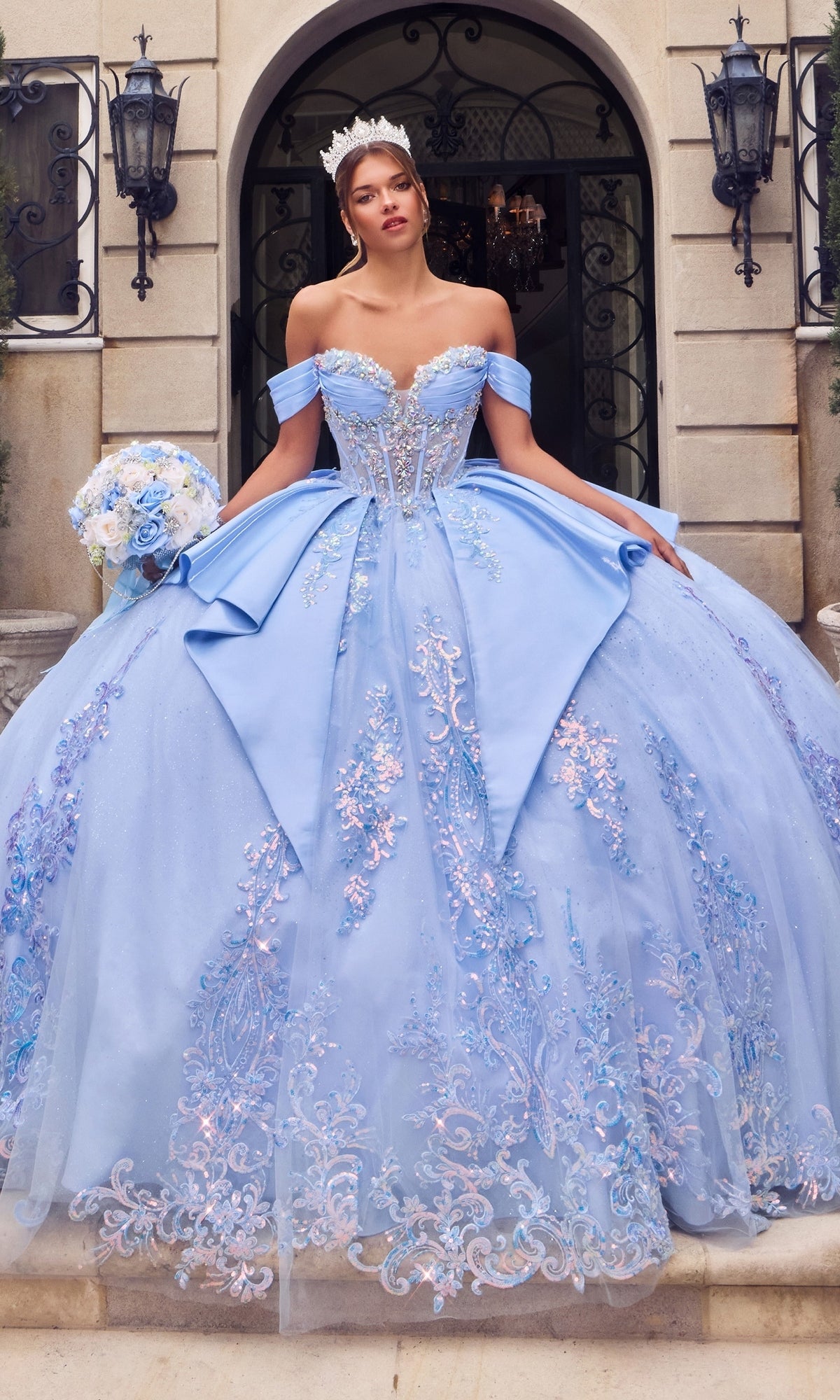 A woman wearing the Quinceanera Dress 15745 by Ladivine in light blue, featuring a glitter tulle skirt and floral details, stands on stone steps holding a bouquet and wearing a tiara in front of an ornate building entrance.